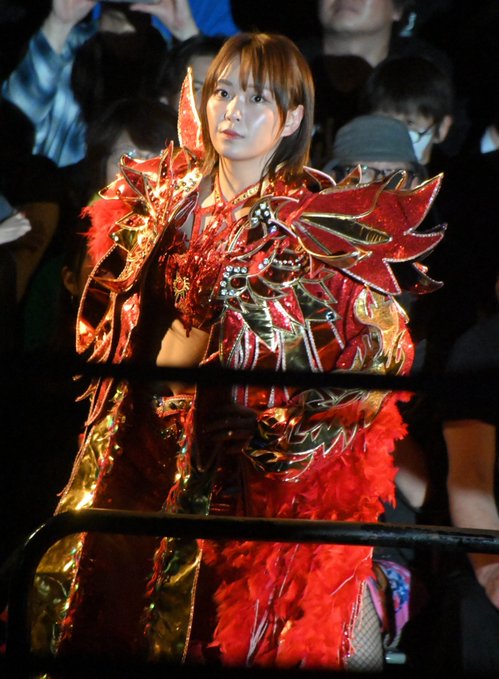 A female professional wrestler wearing an ornate red and gold feathered costume with spiked shoulder pieces stands in a wrestling ring.