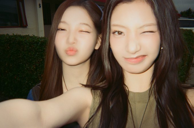 Two young women take a selfie together outdoors, both winking and making playful kissing expressions at the camera.