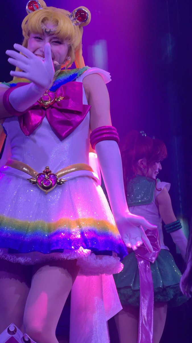 A cosplayer dressed as Super Sailor Moon in a colorful rainbow-hemmed sailor fuku performs on stage under vibrant purple and pink lighting.