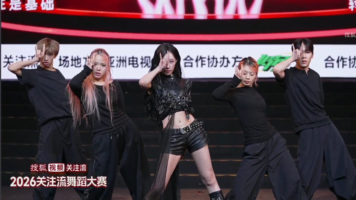 The post shows three performers in matching black outfits striking a choreographed dance pose with two male dancers flanking them, with the central performer featuring long dark hair and a black leather harness outfit. The image relates to the post by showcasing the performer Handong, as indicated by the hashtag, in an intense dance moment at the "2026关注流舞蹈大赛" (2026 GuanZhuliu Dance Competition), with her hand raised dramatically near her face in a fierce expression.