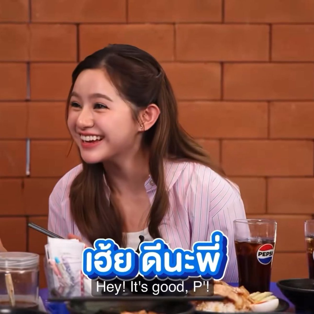 A smiling woman in a pink striped shirt sits at a table with Pepsi drinks and food, with Thai subtitle 'เฮ้ย ดีนะพี่' and English translation 'Hey! It's good, P'!'
