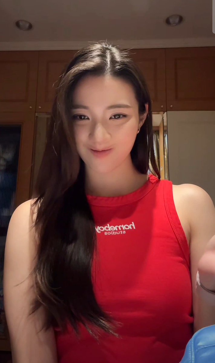 A young woman with long dark hair smiles at the camera while wearing a red sleeveless shirt with mirrored 'noitarepo estime' (Estimation Operation) text on the chest, likely taken as a selfie in a home setting.