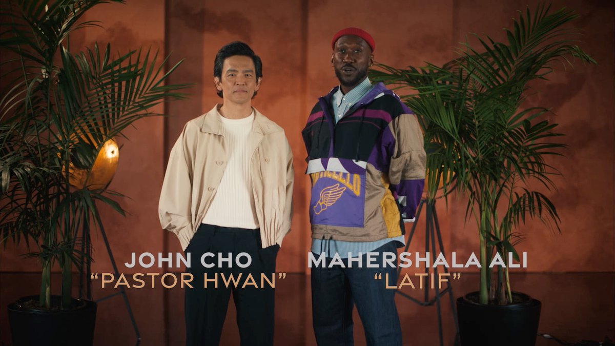 John Cho as "Pastor Hwan" and Mahershala Ali as "Latif" stand side by side in a promotional still for the film "Your Mother," with on-screen text identifying the actors and their characters against a warm backdrop flanked by potted palms.