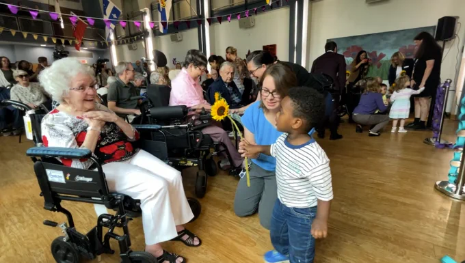 Young boy offering a sunflower to a smiling Senior woman in a wheelchair during a lively community event indoors.