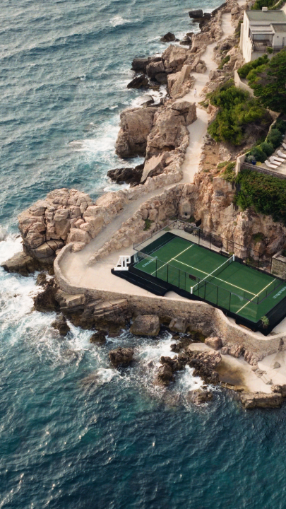 An aerial view of a coastal property featuring a green tennis court and a paddle court built on rocky cliffs next to a building, overlooking the churning blue ocean.