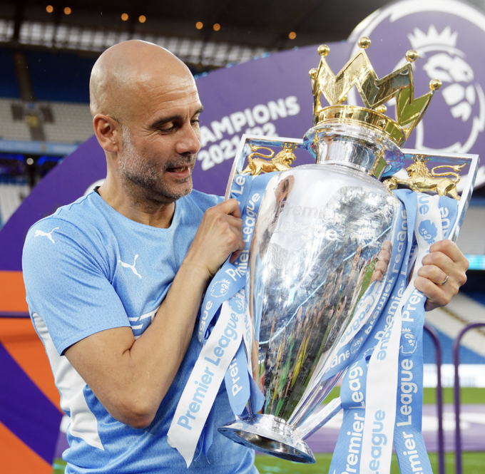 Pep Guardiola, manager of Manchester City, gazes down at the Premier League trophy with a slight smile, the "Premier League" branded ribbons draped across the silver cup. In the background, a banner reads "CHAMPIONS 2021/22," highlighting one of the title wins that could now be at risk under the threat of retrospective punishment.