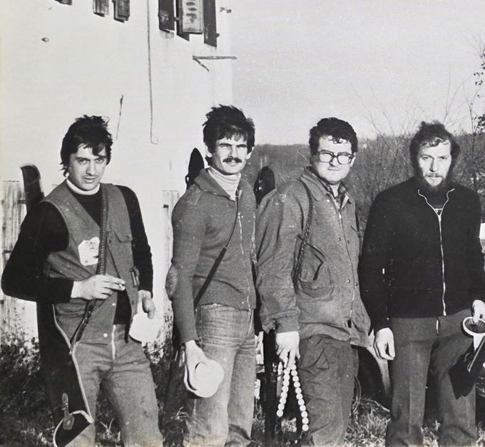Four men stand outdoors in black and white, dressed in casual jackets and vests, holding what appear to be skis, with a building and bare trees visible in the background. The image appears to capture the photographer's father and his hunter friends from Mercato Vecchio, a fraction of Montebelluna, in 1981.