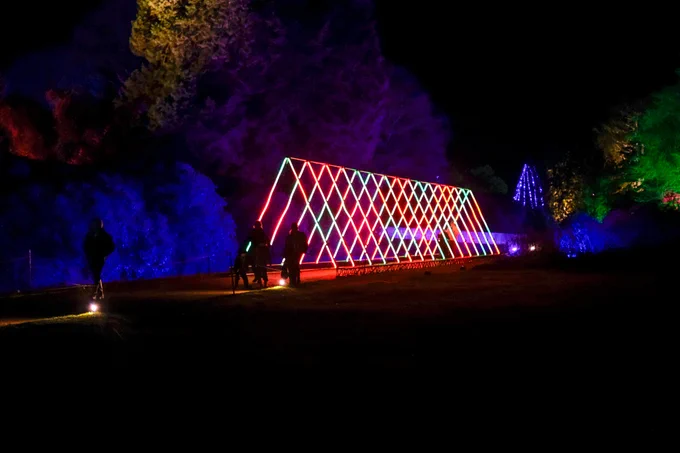 A nighttime scene showcasing a large, illuminated triangular tunnel structure with vibrant red and green lights. The installation is set in a park with trees and bushes in the background, also lit in various colors. Silhouettes of people are visible near the installation.
