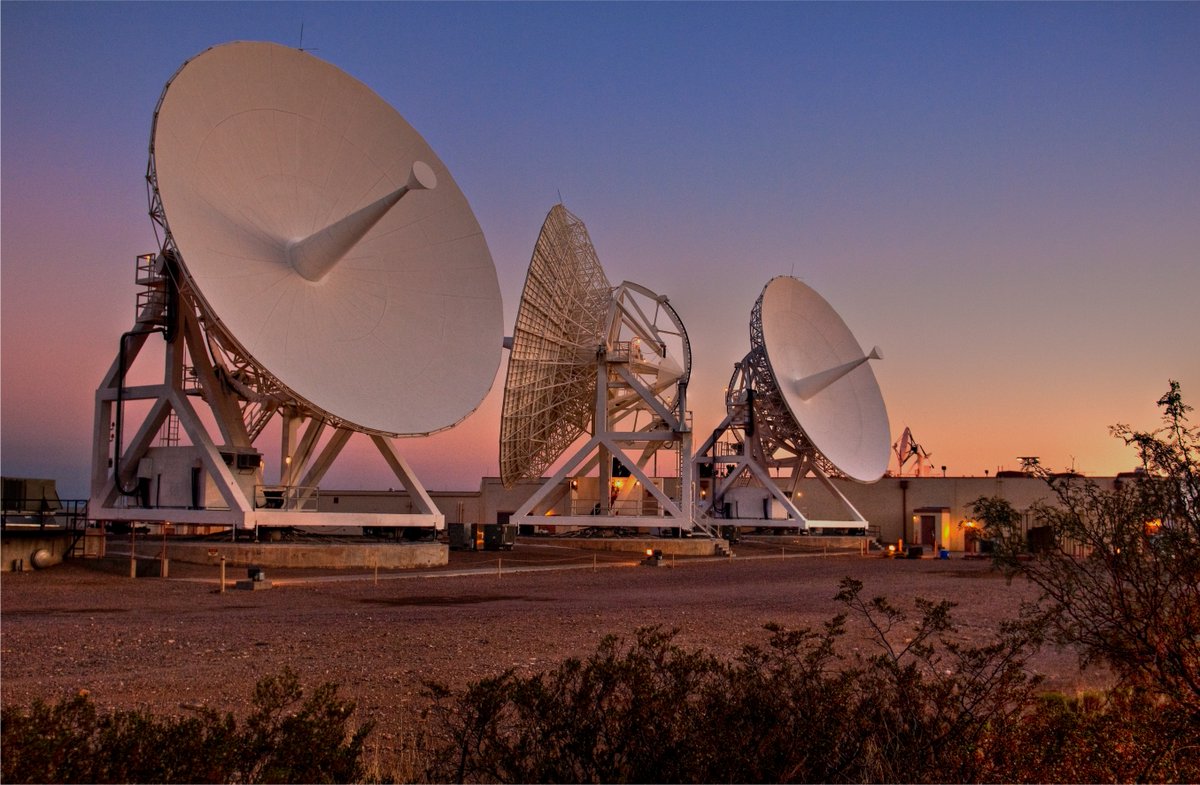 Three large satellite dishes stand outdoors on metal supports, angled toward the sky during sunset. The ground is covered in gravel, a few shrubs sit in the foreground, and the sky fades from warm orange near the horizon to deep blue above.