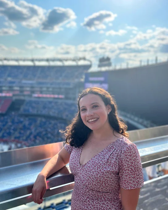 Sara smiles in front of a stadium, with the field and stands behind her.