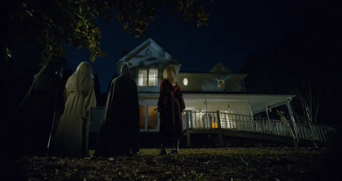 A still from the film The Heretiks. Four women in long robes can be seen from behind, at night, standing in a line and looking at a suburban house.