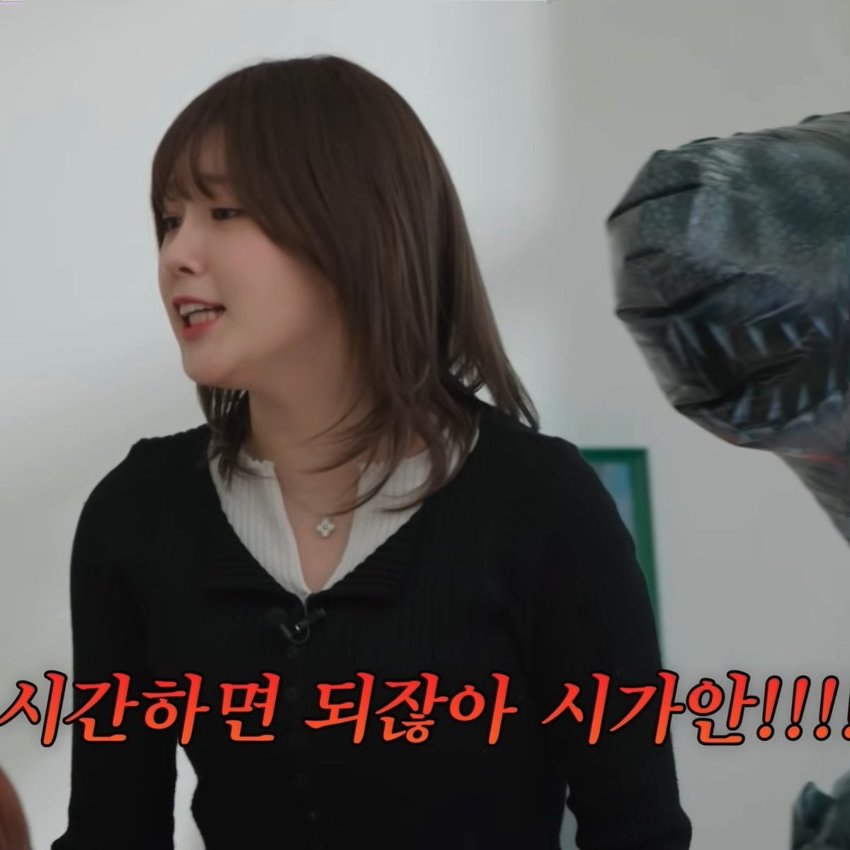 A woman with brown hair wearing a black cardigan over a white shirt speaks in an indoor setting, with a dark mask partially visible on the right and orange Korean text at the bottom reading '시간하면 되잖아 시간!!!'.
