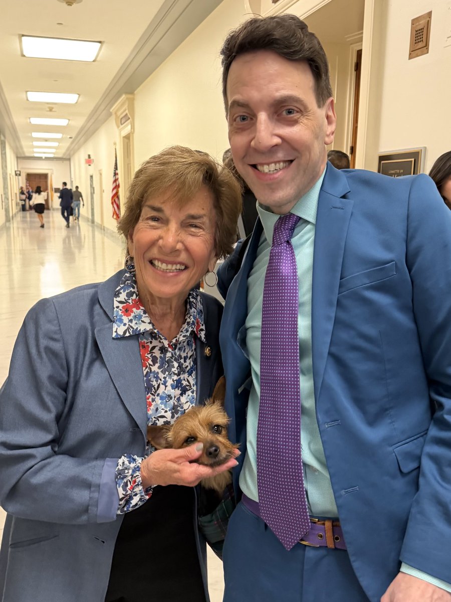 A smiling older woman and a smiling man in a blue suit pose together in a hallway. The woman is holding a small dog. Other people and an American flag are visible in the background. There is some text on a wall plaque.