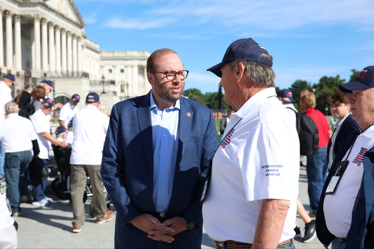 An outdoor gathering near a large classical building. A man in a blue suit and glasses talks with an older man wearing a cap and a white veterans' shirt, while other people stand nearby. Text is visible on some clothing and caps.