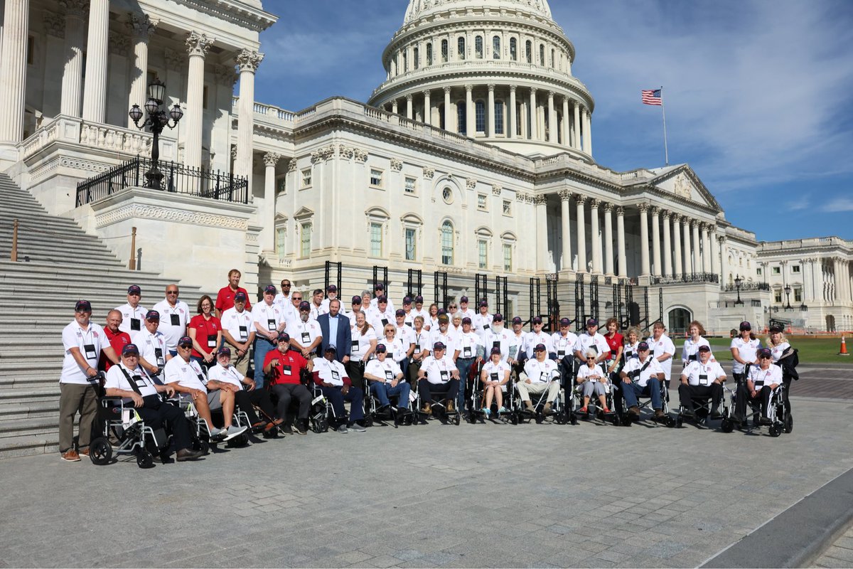 A large group of people, many using wheelchairs, pose for a photograph in front of the United States Capitol on a sunny day. An American flag flies above the building. No visible text.