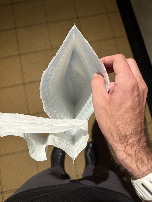 A hairy hand holds open the empty interior of a clear plastic baggie, revealing its diamond-shaped opening, over a tiled floor. The post humorously rebrands the empty plastic bag as a "packagussy," playing on the visual of the bag looking like an empty wrapper.