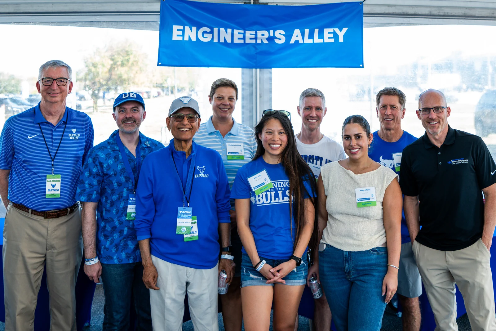 Adults (6 men, 3 women) smile at the camera under a blue 'ENGINEER'S ALLEY' banner. Most wear blue shirts, some with 'Buffalo Bulls' or 'UB' logos.