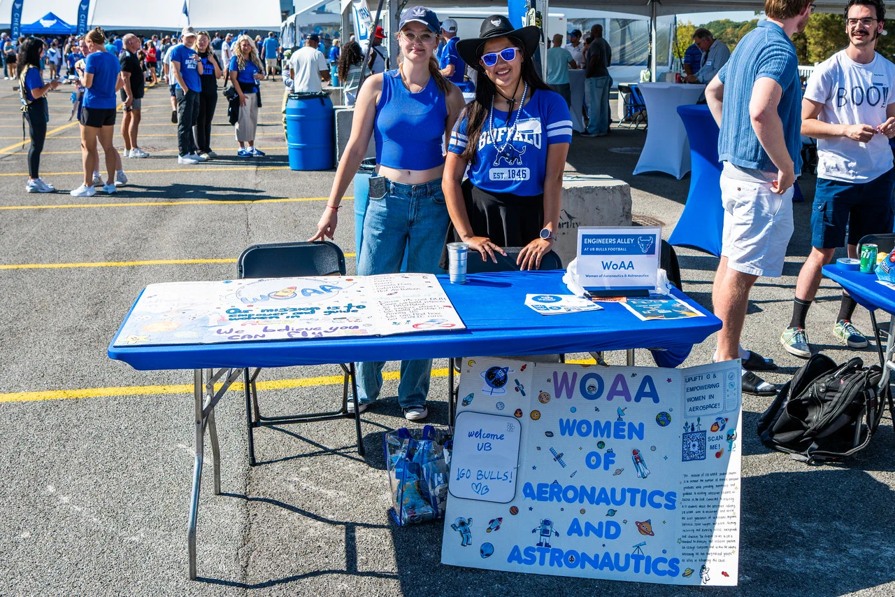 Two women, one in a cowboy hat and sunglasses, smile behind a blue table at an outdoor event. 