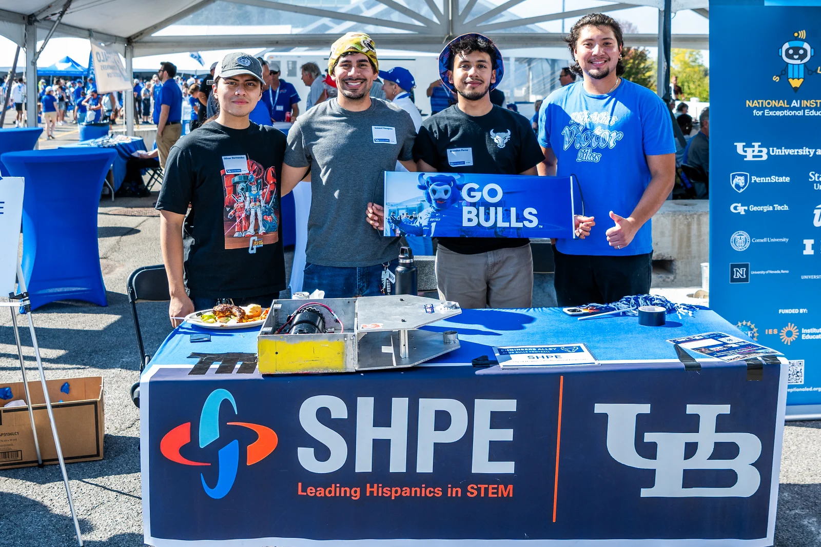 Four smiling young men stand behind a table at an outdoor event. They hold a blue banner that reads \"GO BULLS\". 