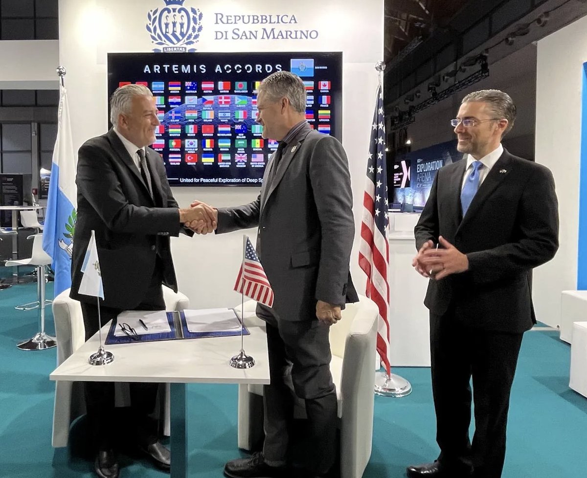 Two men shake hands while standing over signed papers on a desk. A TV screen in the background displays the flags of the Artemis Accords nations. A man stands close by and smiles.