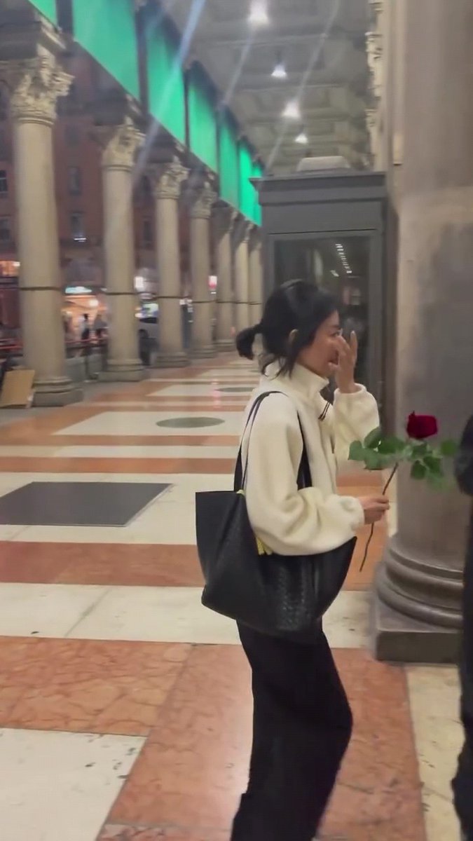 A young woman with dark hair tied back walks through a grand columned arcade, wearing a cream-colored fleece jacket and carrying a black tote bag while holding a red rose and touching her face.