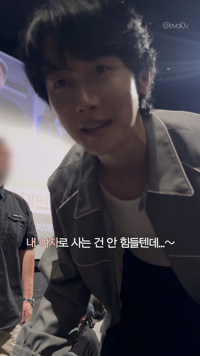 Close-up photo of a young man with curly black hair in a gray jacket, with Korean text overlay reading '내 여자로 사는 건 안 힘들던데...~' and the handle '@evol.OV'.