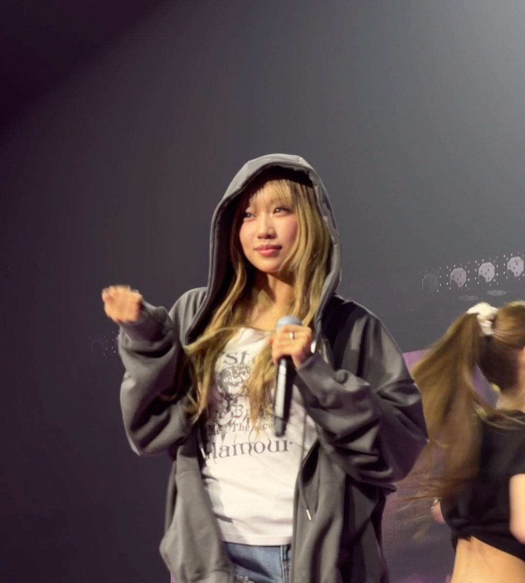 A concert photo of a young woman with blonde-streaked hair wearing a grey hoodie over a graphic t-shirt, holding a microphone while performing on a dimly lit stage.