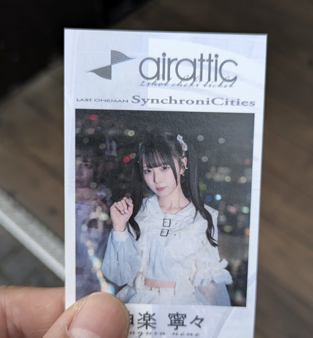 A hand holds a trading card or photocard featuring a young woman in a white dress against a city night background, labeled 'airattic LAST ONEMAN Synchronicities' with Japanese characters below.