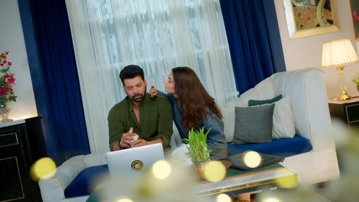 A man and a woman sit together on a couch looking at a laptop; the woman leans in to whisper to the man while he holds a small cup.