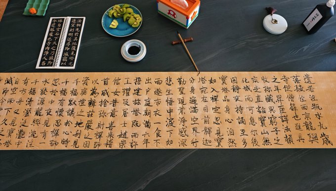 A long scroll of Chinese calligraphy is laid out on a dark surface, surrounded by traditional items including a plate of kiwi slices, a small ink stone, and a wooden stamp.