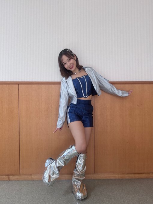 A young woman poses playfully against a wooden wall, wearing a denim crop top and shorts with a silver jacket and distinctive silver metallic leg coverings.