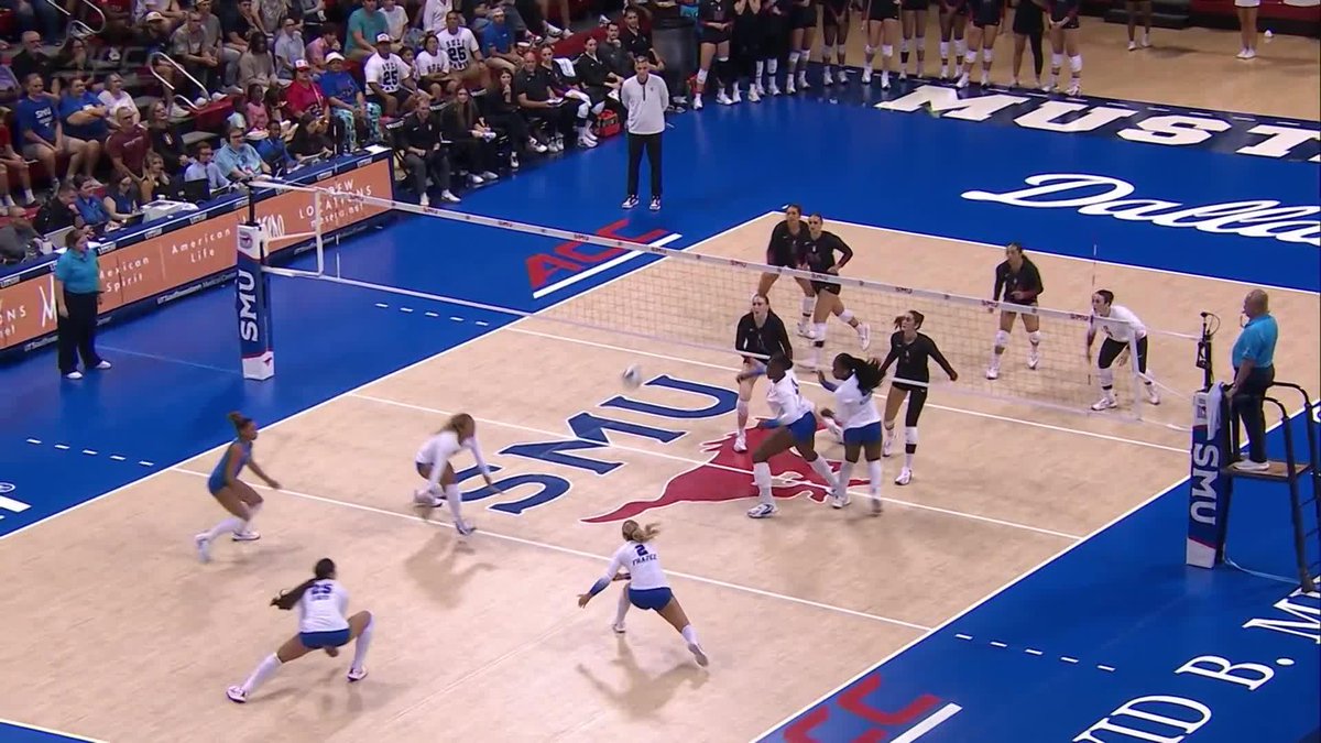 A women's college volleyball match takes place on a court with 'SMU' and a Mustang logo, showing players in action during a game with spectators watching from the sidelines.