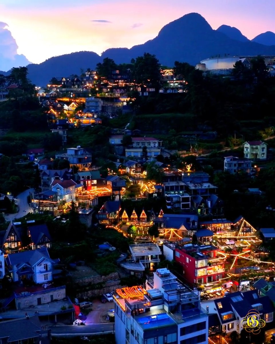 A twilight panoramic view of a hillside town illuminated with countless warm string lights, nestled against a silhouette of jagged mountains under a pink and purple sky.