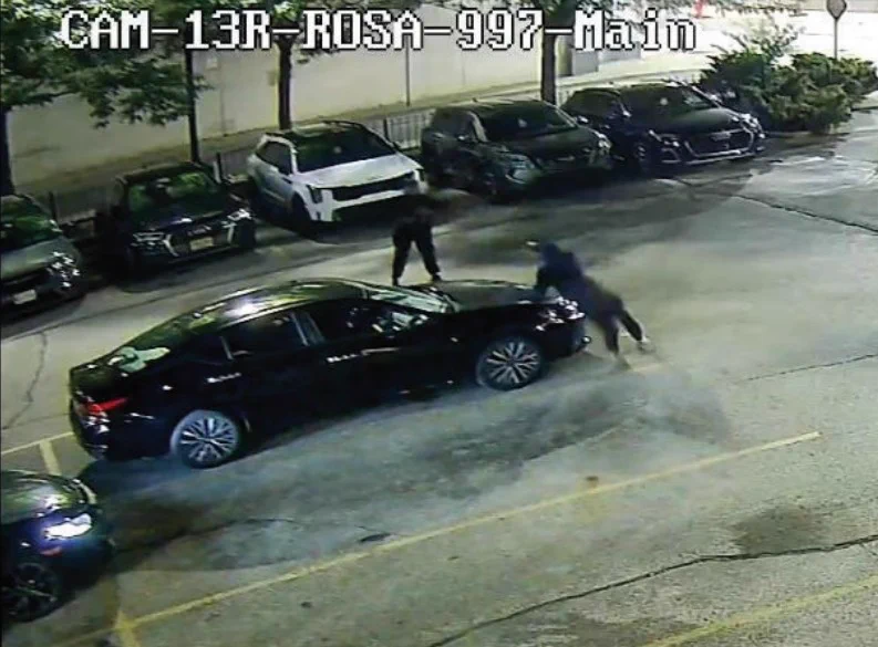 Two individuals appear to be tampering with a black sedan in a dimly lit parking lot at night.