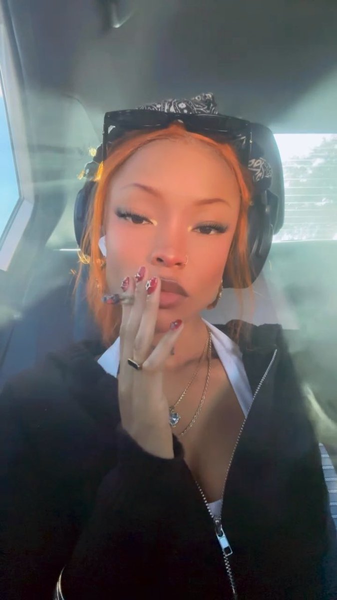 A woman with bright orange hair, sunglasses on her head, and over-ear headphones poses for a selfie while holding a vape, surrounded by vapor.
