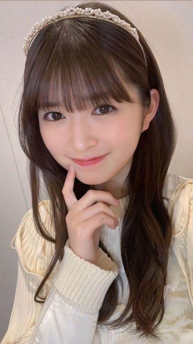 A selfie of a young woman with long brown hair wearing a pearl headband and a cream-colored sweater, smiling with her finger near her chin.