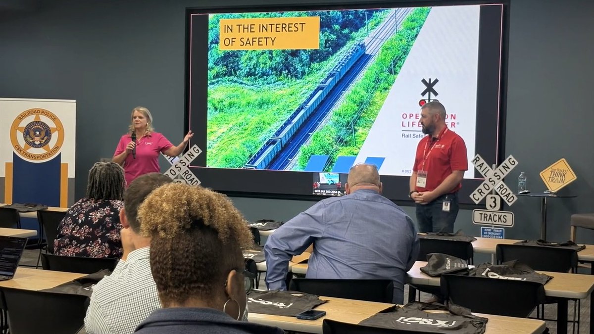 A railroad safety presentation where a woman and a man in red shirts stand before a large screen displaying 'IN THE INTEREST OF SAFETY' and 'OPERATION LIFESAVER', addressing an audience in a classroom setting.