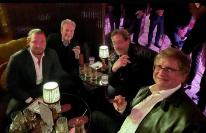 Four men sit around small tables in a dimly lit lounge or bar, drinking and smoking cigars.