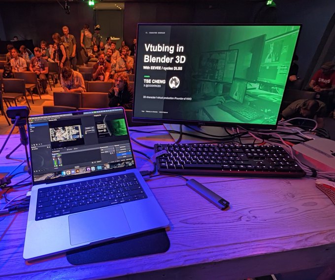 A workspace setup shows a laptop running OBS software next to a monitor displaying a presentation slide titled 'Vtubing in Blender 3D' with 'TSE CHENG' listed as the speaker, set in an auditorium.