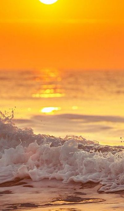 A vertical photograph of ocean waves crashing onto a beach at sunset, with the sun glowing orange on the horizon and reflecting across the water.