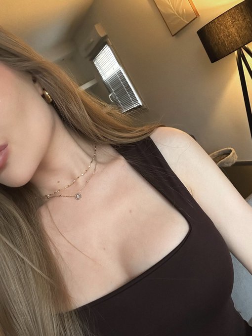 A close-up selfie of a woman wearing a dark top, layered gold necklaces, and earrings, taken indoors near a lamp and window blinds.