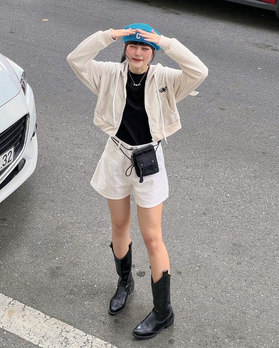 A young woman poses on a street wearing a blue helmet, a beige zip-up jacket, white shorts, a small black waist pouch, and black cowboy boots.