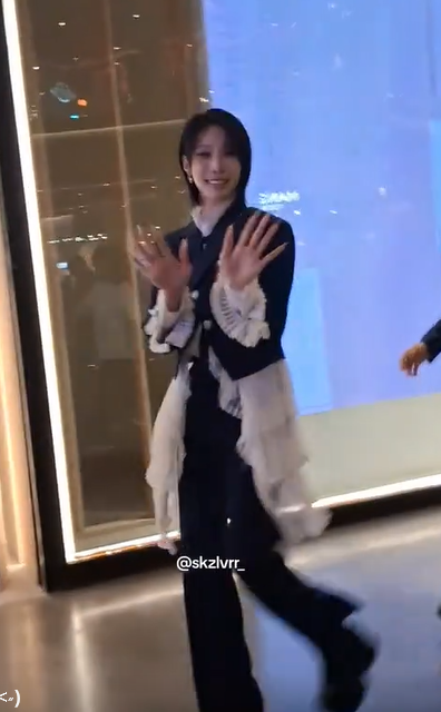A stylish person poses playfully with both hands raised while walking through a brightly lit modern space, wearing a dark top with white ruffled accents, black pants, and boots. The watermark "@skzlvrr_" is visible at the bottom of the frame, matching the fan's excitement about finally catching a glimpse of today's outfit.