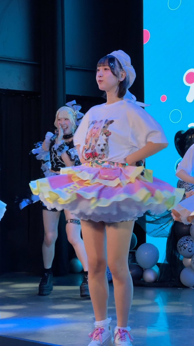 A young woman stands on a brightly lit stage wearing a white t-shirt with an anime print, a colorful pastel ruffled skirt, and a white headscarf. Other performers are visible in the background.