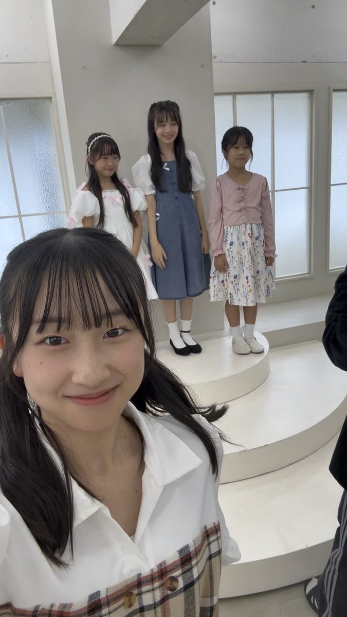 A selfie of a smiling young woman in the foreground with three other young girls standing on a tiered white platform in the background, all dressed in coordinated outfits.