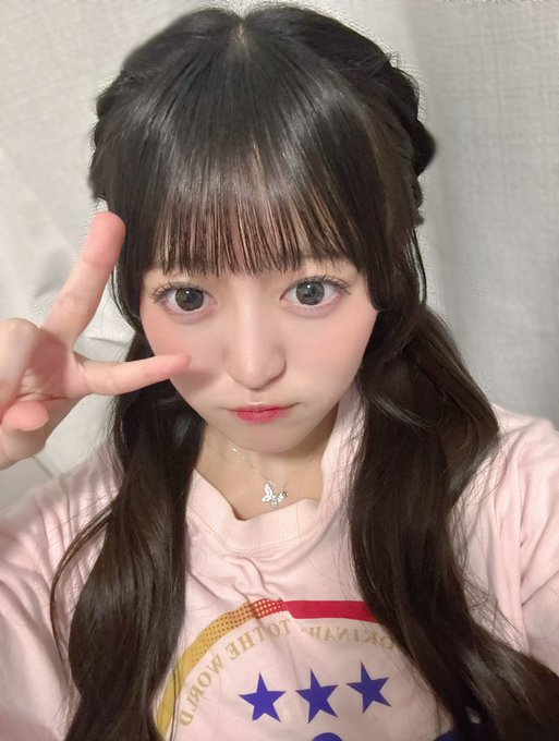 A close-up selfie of a young woman with long dark hair and bangs making a peace sign, wearing a light pink top with star designs.