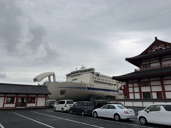 A large white passenger ship docked beside a traditional Japanese building with a tiled roof under a cloudy sky, with several cars parked in the foreground lot.