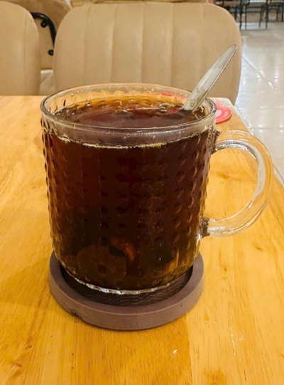 A glass mug filled with a dark iced beverage and ice sits on a wooden table with a spoon inside.