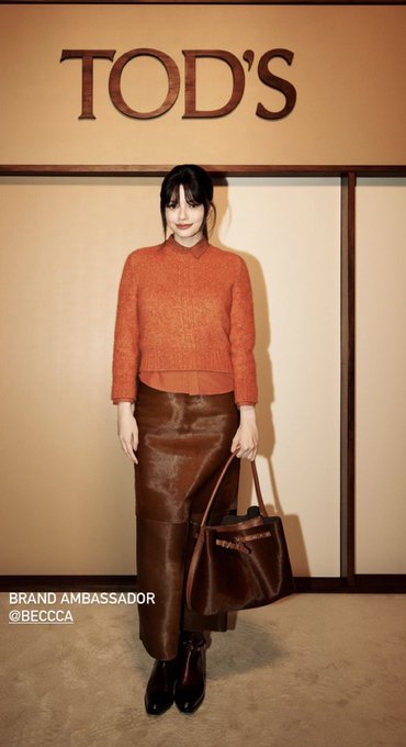 A woman stands in front of a wall featuring the 'TOD'S' logo, wearing an orange sweater, a brown leather skirt, and carrying a matching tote bag; text on the screen reads 'BRAND AMBASSADOR @BECCCA'.