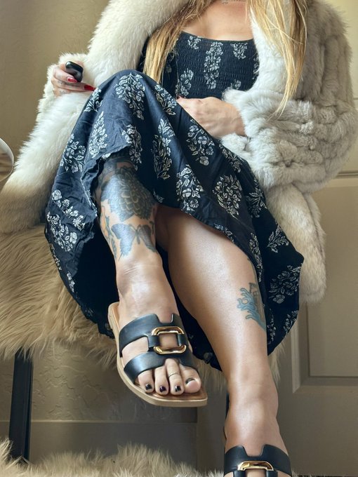 Close-up photo showing a woman's legs with visible tattoos wearing black sandals and a floral dress, sitting on what appears to be a fur seat.